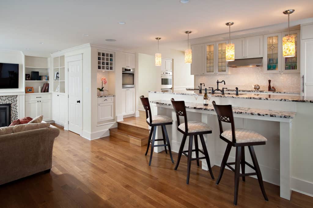 Queens after renovation, a white kitchen and granite island with stools opening to the living room.