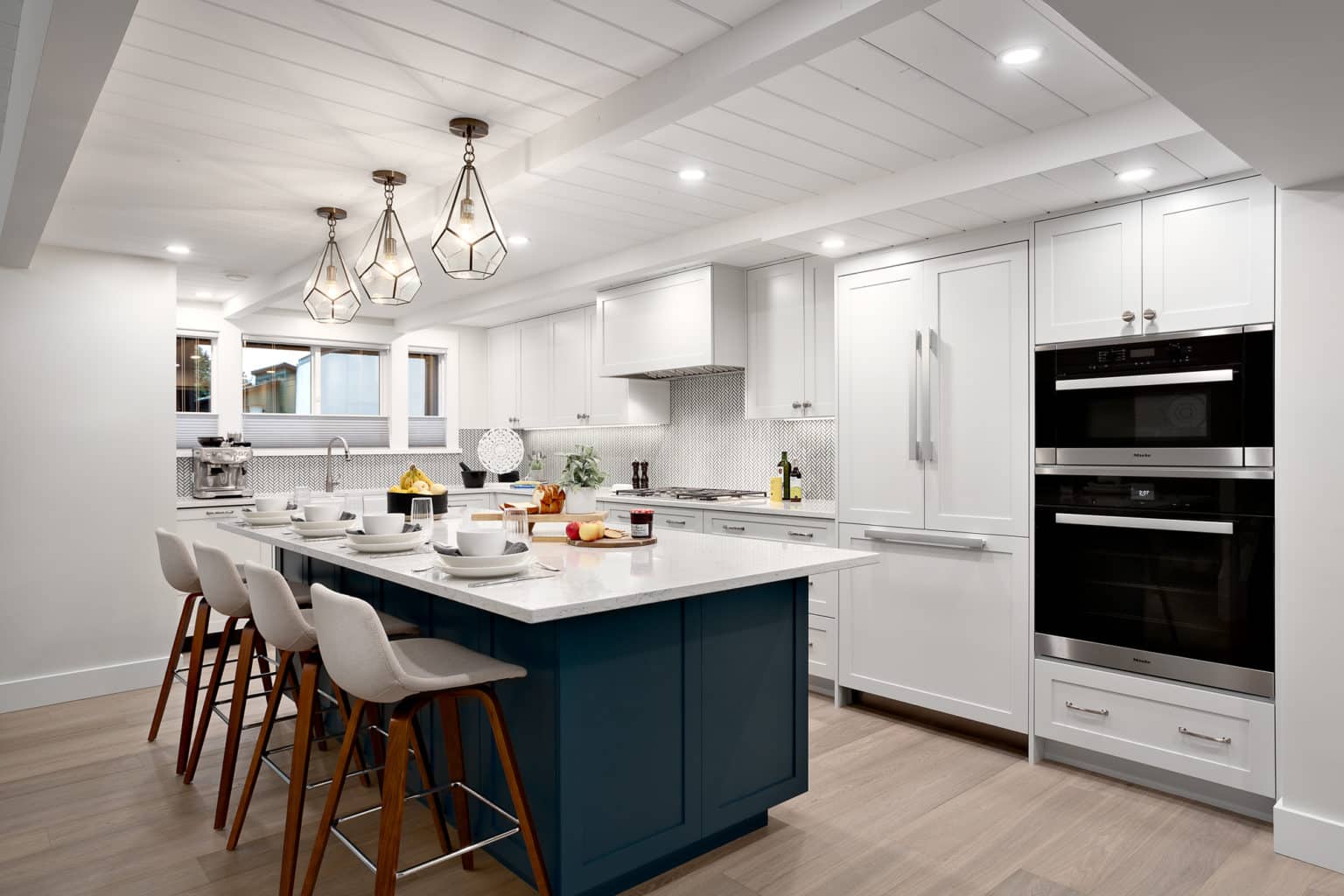 White kitchen with a navy island, three glass pendant lights, counter stools and stacked wall ovens