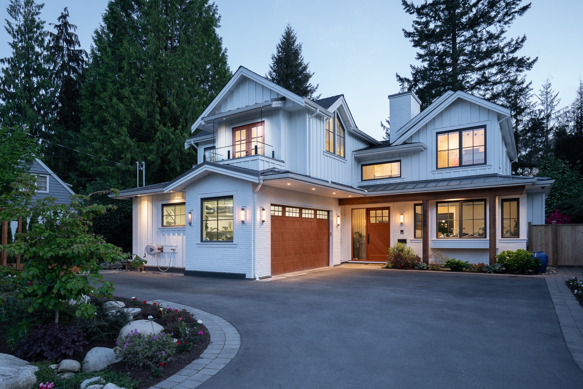 White board-and-batten custom home with a cedar timber porch, lit windows and spring planting at dusk, North Vancouver