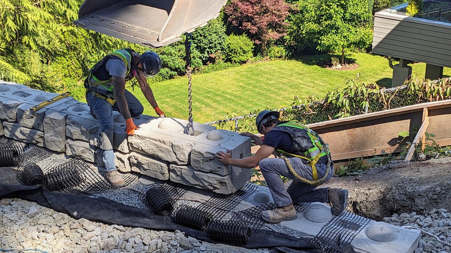 A Shakespeare Homes crew member guiding a lock block into place on a retaining wall, with geogrid and drainage laid out beneath it