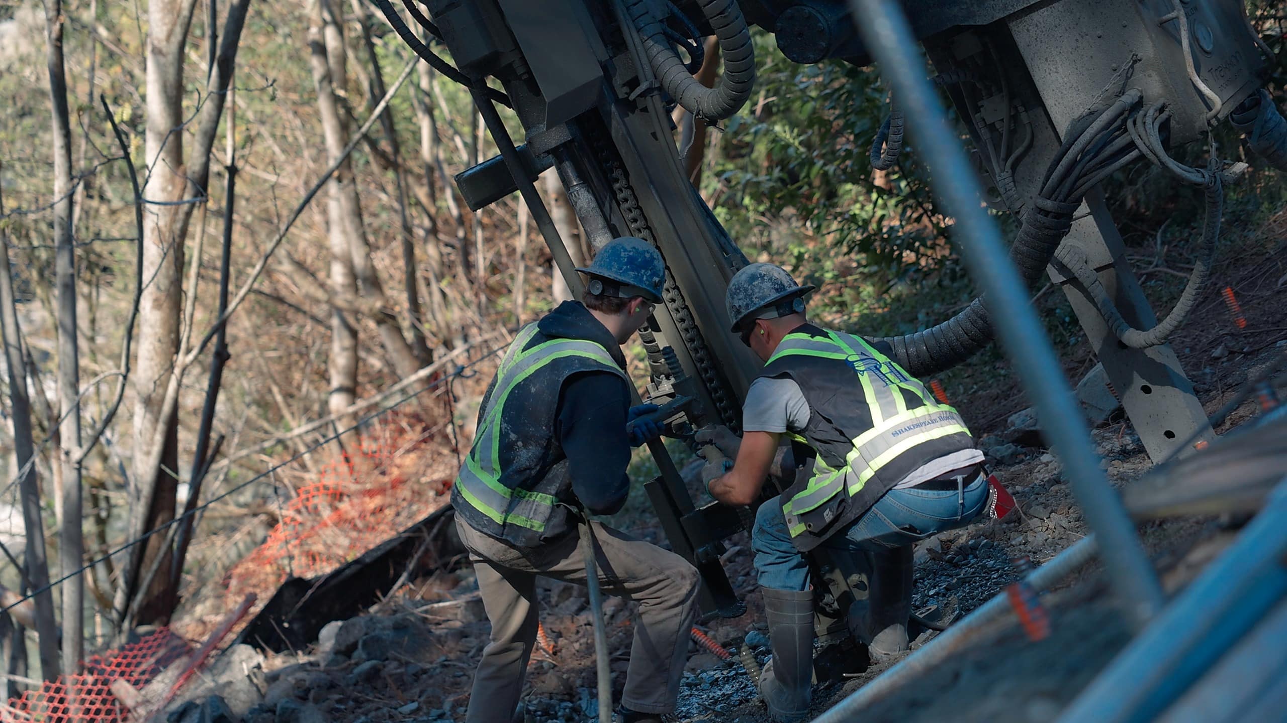 Two Shakespeare Homes crew members working a rock drill on a steep forested slope at the Isleview site