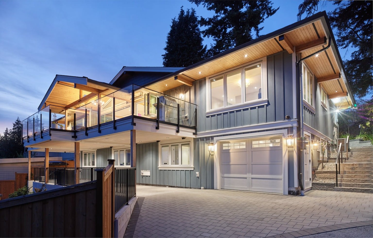 A two-storey West Coast contemporary home lit from within at dusk, its sage-green board-and-batten siding and cedar-lined soffits framing a glass-railed deck above a covered patio, with a paver driveway running to the double garage.