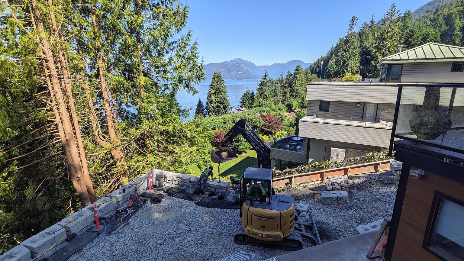 An excavator working a constrained hillside lot between two houses above Howe Sound, with a block retaining wall going in