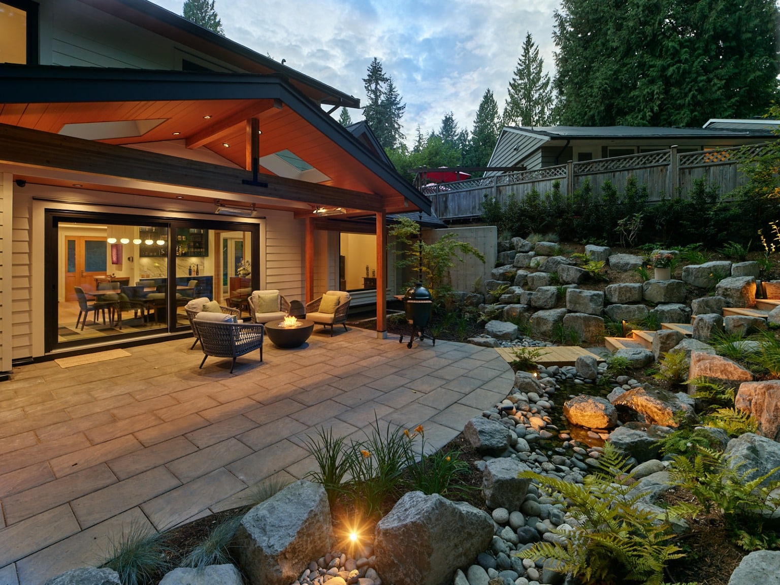 Terraced boulder retaining wall with fern and grass planting beside a covered patio and fire bowl, lit at dusk