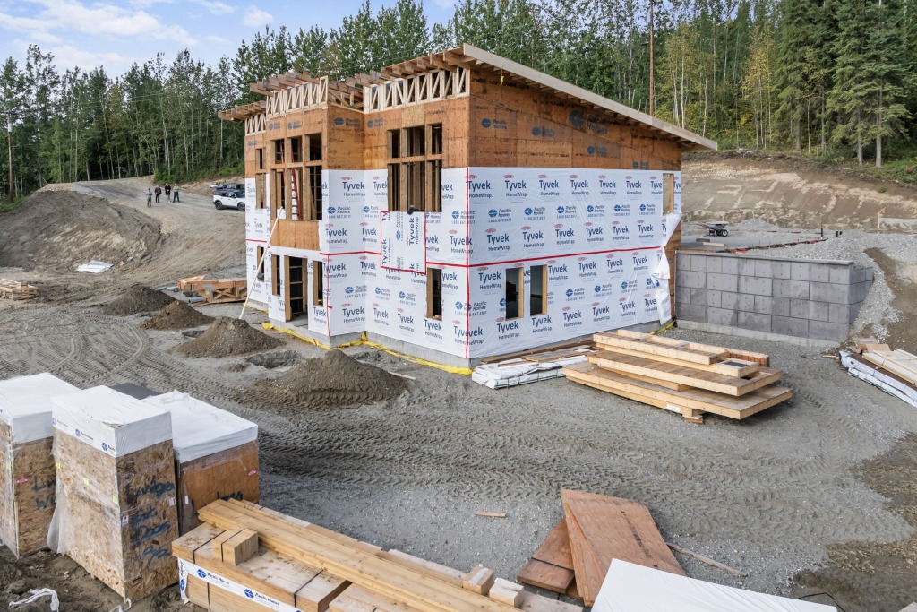 A prefabricated house frame wrapped and standing on its foundation, with panel packages waiting on site