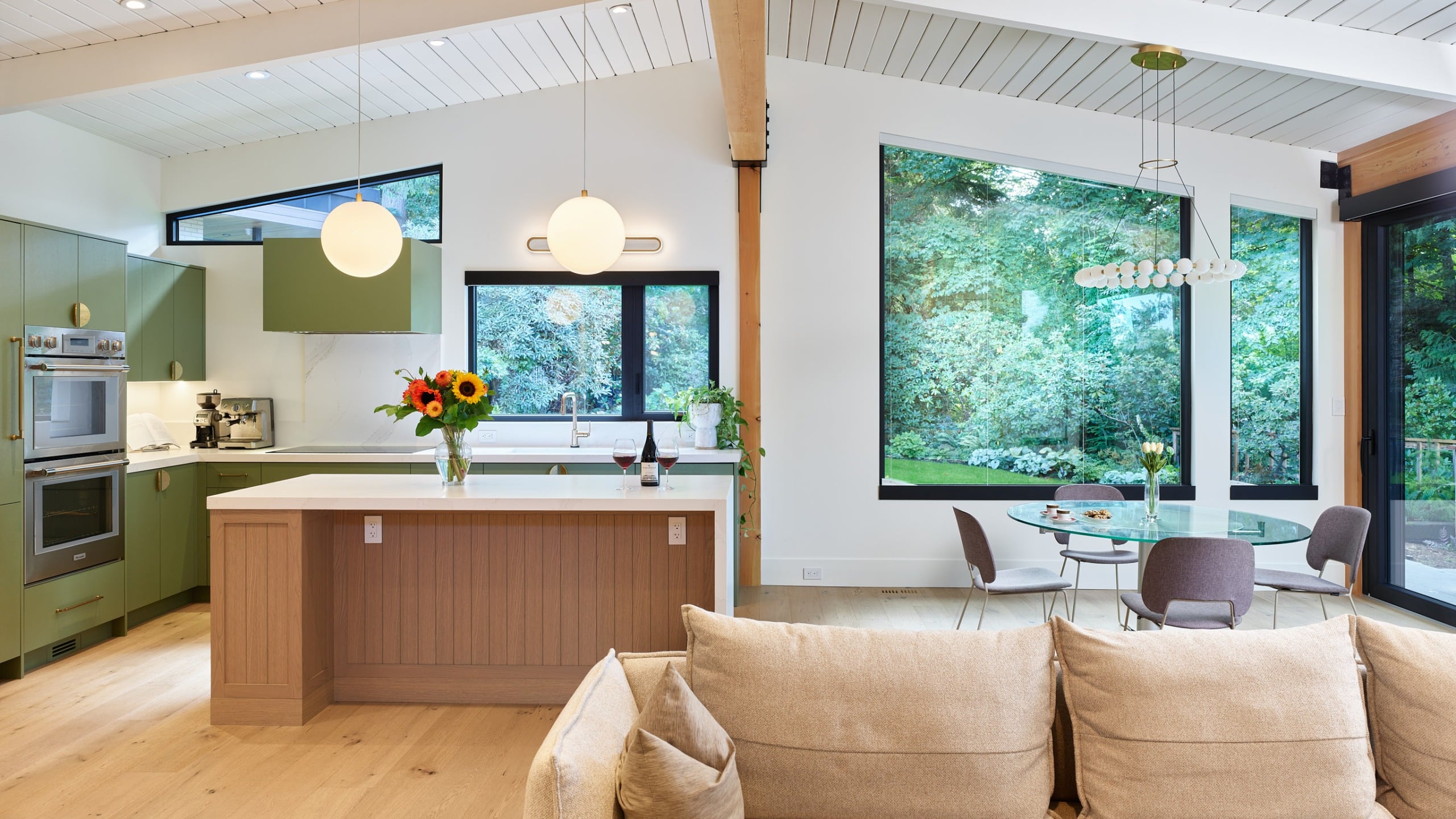 Window Into The Woods, an open plan kitchen and living room with sage green cabinetry, a pale oak island and a vaulted plank ceiling, open to the dining room and the garden, North Vancouver